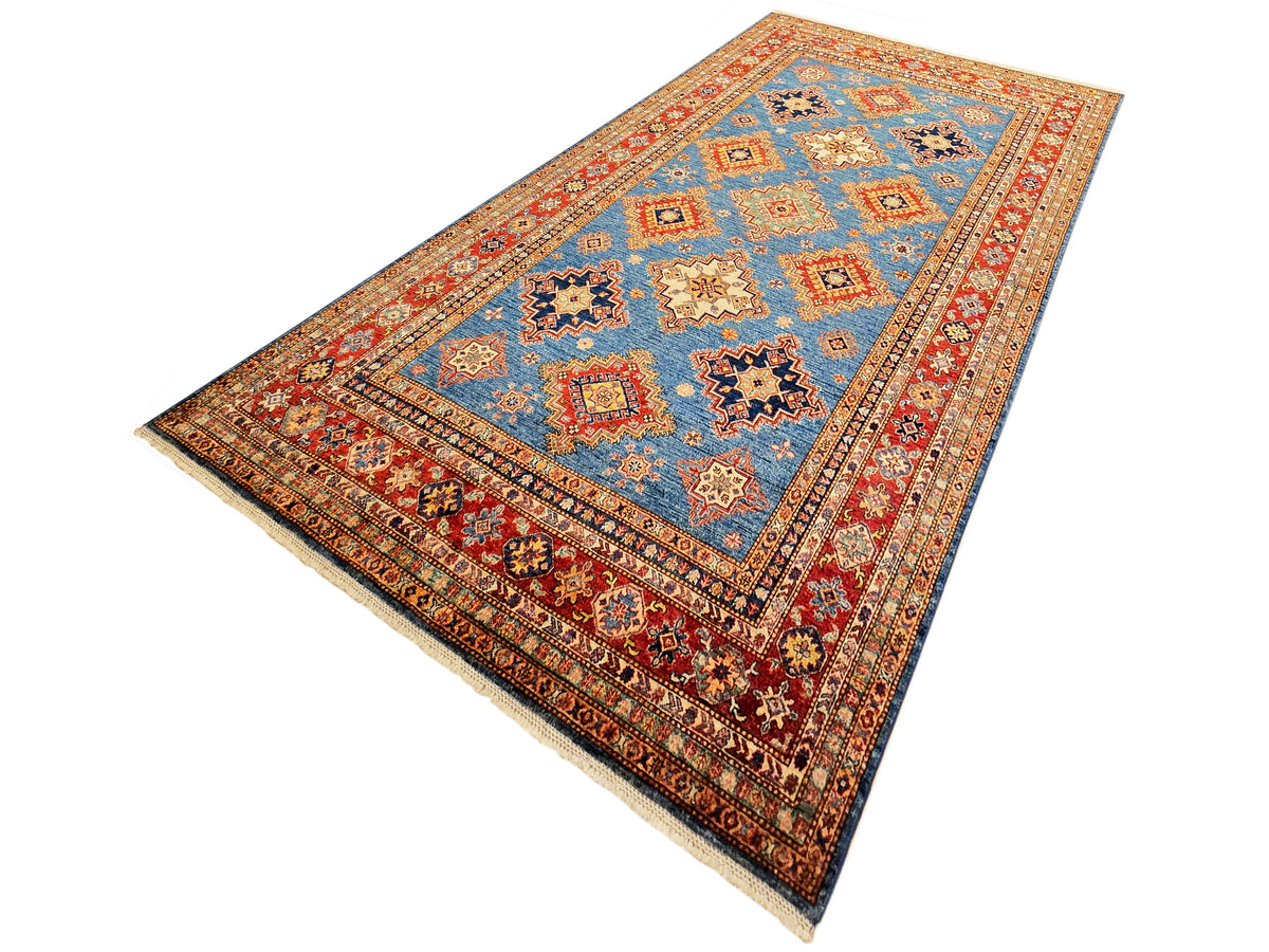 Angled view of geometric blue rug with ornate red border and diamond motifs.