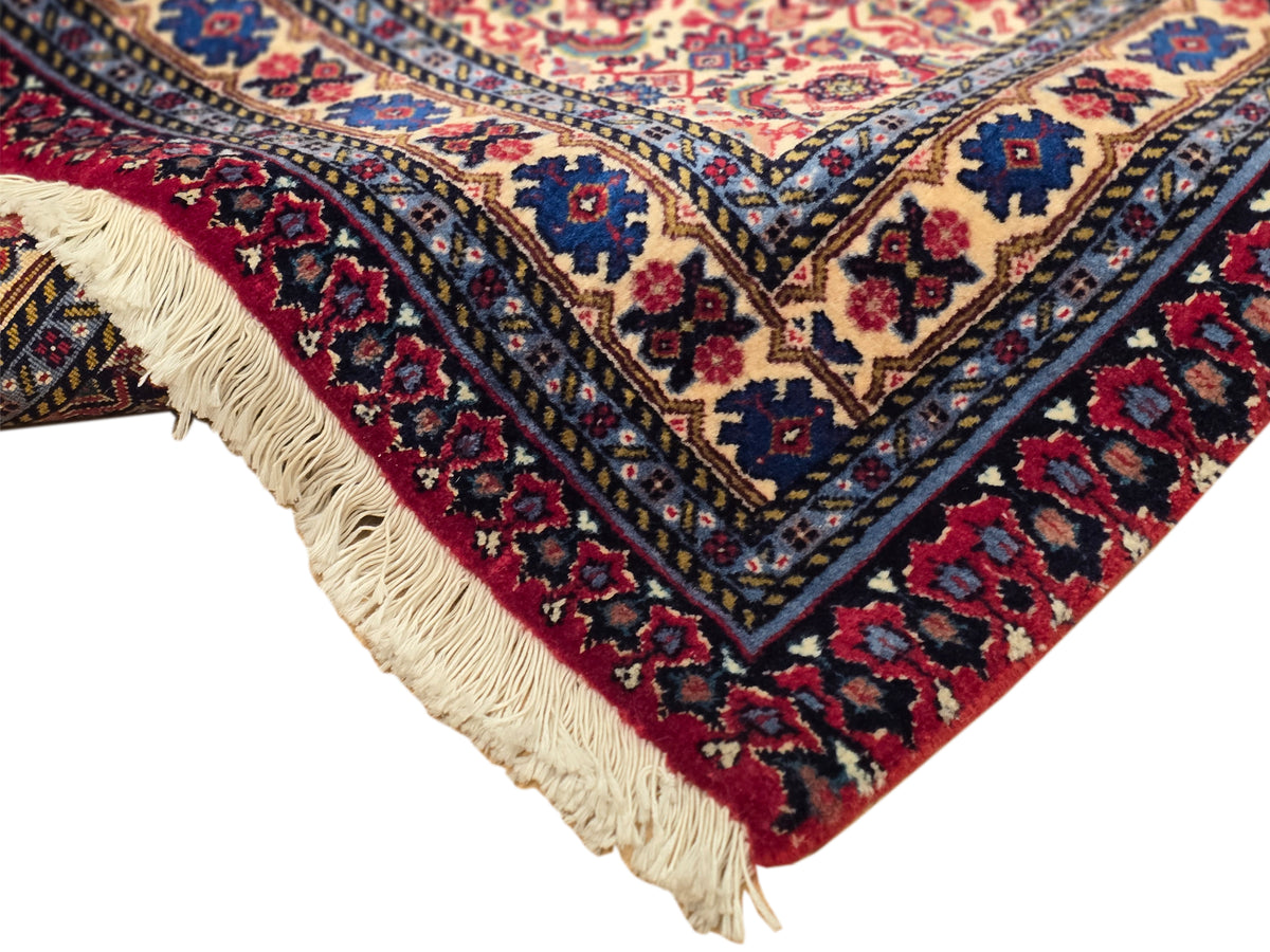 Corner of patterned area rug with red field, navy trim, and cream fringe tassels.