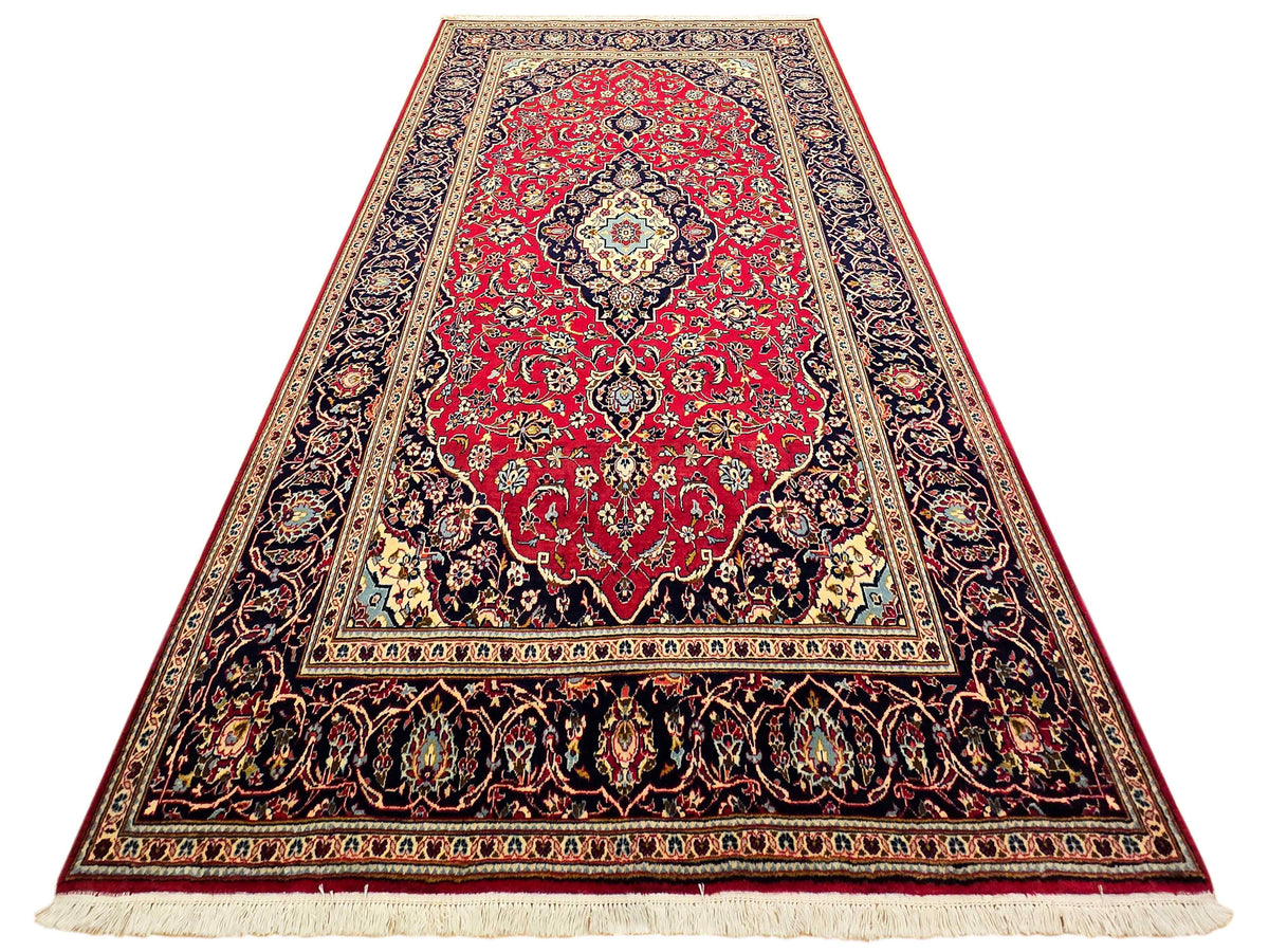 Kashan rug on white background, red medallion with navy border and cream fringe